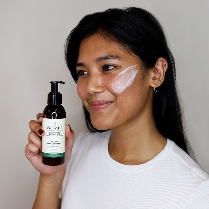 Woman holding a bottle of Sukin facial cleanser with product on her face.