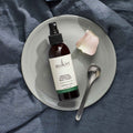 Sukin bottle on a gray plate with a rose petal and spoon on a blue fabric background